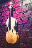 A violin is mounted against a textured brick wall painted in purple hues, with strings crisscrossing the wall and small pegs attached to them.