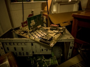 A partially disassembled computer tower with various circuit boards and electronic components. The setup is cluttered, with a cardboard box and table nearby. The environment seems dimly lit, emphasizing the worn and outdated technology.
