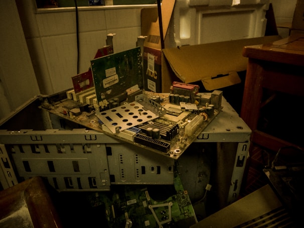 A partially disassembled computer tower with various circuit boards and electronic components. The setup is cluttered, with a cardboard box and table nearby. The environment seems dimly lit, emphasizing the worn and outdated technology.