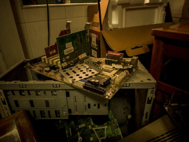 A partially disassembled computer tower with various circuit boards and electronic components. The setup is cluttered, with a cardboard box and table nearby. The environment seems dimly lit, emphasizing the worn and outdated technology.