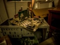 A partially disassembled computer tower with various circuit boards and electronic components. The setup is cluttered, with a cardboard box and table nearby. The environment seems dimly lit, emphasizing the worn and outdated technology.