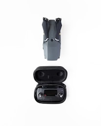 A folded grey drone is placed above a black case containing a remote controller, both set against a white background.