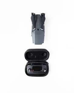 A folded grey drone is placed above a black case containing a remote controller, both set against a white background.