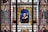 A detailed stained glass window depicts various figures associated with Shakespeare's works. In the center is a portrait of Shakespeare himself, surrounded by colorful, intricate patterns and designs. On either side are characters from his plays, including Juliet and Lady Macbeth. The artwork is rich in symbolism and ornate detail, conveying a sense of grandeur and reverence for the literary figures.