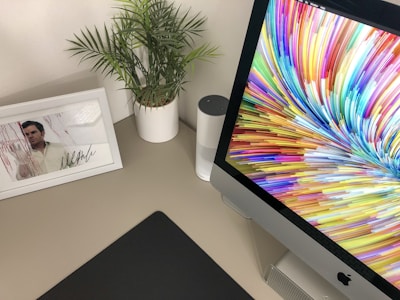 A desk setup featuring a computer display with a vibrant, colorful screensaver featuring a swirling pattern. To the left, there is a small potted plant with green leaves and a framed photograph of a person with a signature underneath. A cylindrical smart speaker can be seen partially obscured behind the plant.