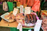 A market stall displaying an assortment of dried foods, including fish, sausages, and other meats. Handwritten signs with prices are visible, written in Chinese characters. The stall background includes green elements and various hanging items, contributing to a bustling market atmosphere.