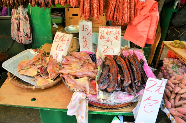 A market stall displaying an assortment of dried foods, including fish, sausages, and other meats. Handwritten signs with prices are visible, written in Chinese characters. The stall background includes green elements and various hanging items, contributing to a bustling market atmosphere.