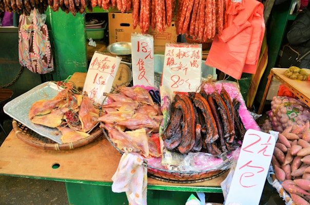 A market stall displaying an assortment of dried foods, including fish, sausages, and other meats. Handwritten signs with prices are visible, written in Chinese characters. The stall background includes green elements and various hanging items, contributing to a bustling market atmosphere.