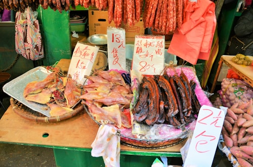 A market stall displaying an assortment of dried foods, including fish, sausages, and other meats. Handwritten signs with prices are visible, written in Chinese characters. The stall background includes green elements and various hanging items, contributing to a bustling market atmosphere.
