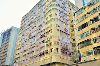 A tall, densely packed residential building with multiple floors in a vibrant urban setting. The building features a facade with a mix of pastel purple and cream colors, numerous air conditioning units, balconies with laundry, and signs written in an Asian script. The architecture exhibits a slightly worn and aged look.
