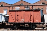 A vintage freight train car is situated on railway tracks in front of a brick industrial building. The train car is primarily red with some weathered textures and markings. The building behind features large glass windows and rounded architectural elements.