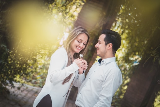A couple is standing close together in a garden-like setting, holding a small flower. Both are smiling warmly at each other, creating a romantic and intimate atmosphere. They are dressed in white shirts, and the background is filled with lush green foliage and soft lighting.