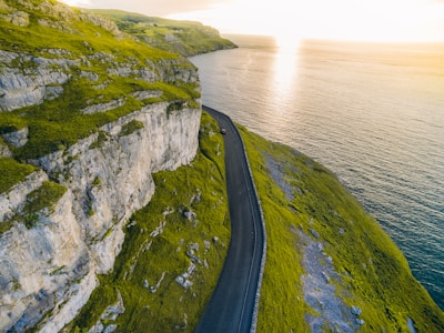 A winding coastal road with waves crashing against the cliffs under a golden sunset.