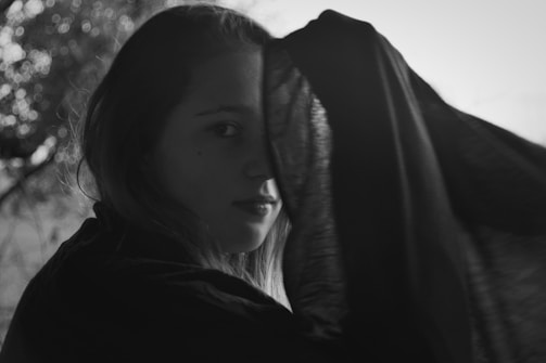 A person is partially obscured by a dark fabric draped over their head, with light softly illuminating their face. The background is blurred, creating a dreamy and intimate atmosphere.