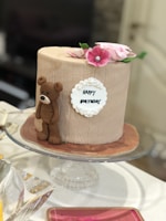 A round, beige birthday cake with a textured surface is adorned with a small, brown fondant teddy bear on its side. Pink and white flowers are arranged on top, along with a white plaque bearing the words 'Happy Birthday' in black icing. The cake is placed on a glass stand.