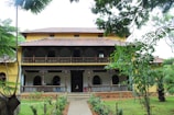 A traditional two-story building with a tiled roof and intricate wooden railings. The facade features arched windows and a spacious veranda. The surroundings are lush with greenery, including several trees and a well-maintained garden.