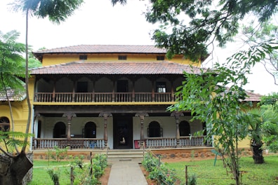 A traditional two-story building with a tiled roof and intricate wooden railings. The facade features arched windows and a spacious veranda. The surroundings are lush with greenery, including several trees and a well-maintained garden.