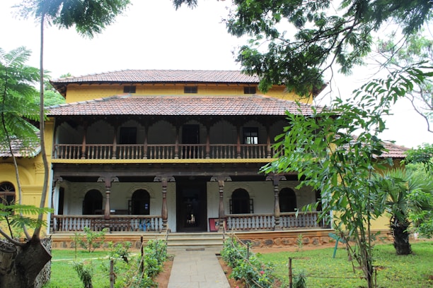 A traditional two-story building with a tiled roof and intricate wooden railings. The facade features arched windows and a spacious veranda. The surroundings are lush with greenery, including several trees and a well-maintained garden.