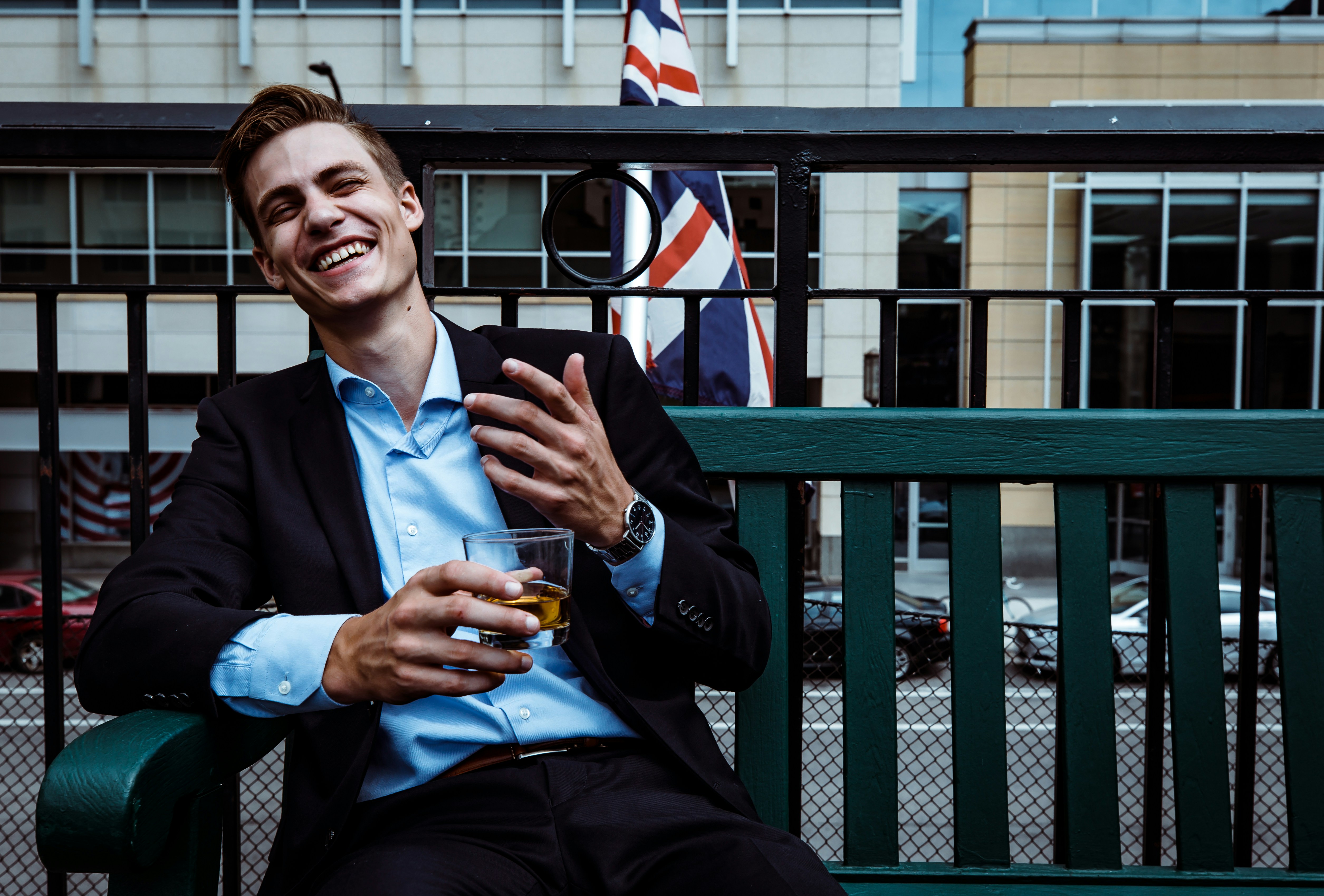man smiling while sitting and holding whisky glass near concrete building, 