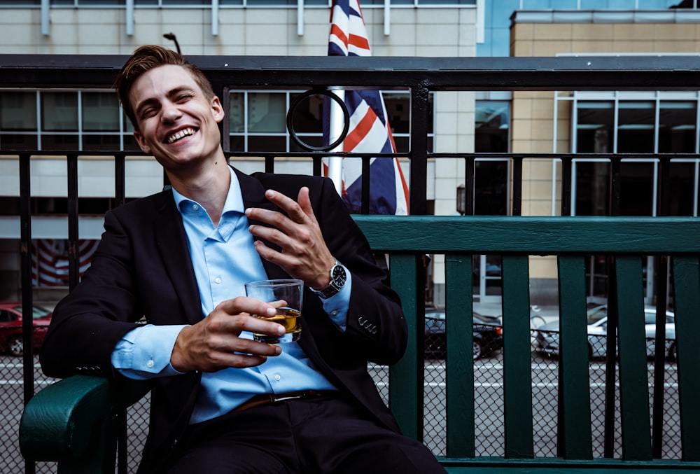 man smiling while sitting and holding whisky glass near concrete building