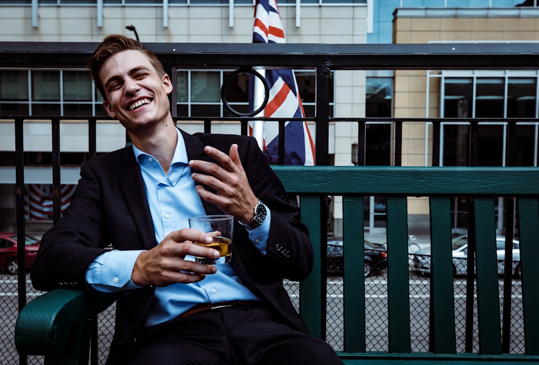 man smiling while sitting and holding whisky glass near concrete building,