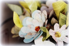 A close-up of intricately crafted paper flowers, with petals made from map and printed paper. The flowers showcase a mix of light blue, yellow, and pink hues on the petals, with green leaves around them. The center of the flowers has detailed rose-like structures, adding depth to the arrangement.