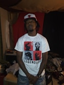 A person wearing a white t-shirt with red and black graphic blocks featuring silhouettes of famous figures, labeled 'Legendary', and a baseball cap with a red bull logo. The person stands with arms down in a dimly lit room with casual furniture and clothing scattered around.