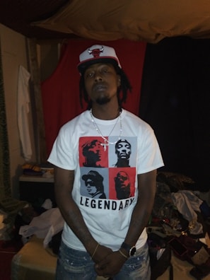 A person wearing a white t-shirt with red and black graphic blocks featuring silhouettes of famous figures, labeled 'Legendary', and a baseball cap with a red bull logo. The person stands with arms down in a dimly lit room with casual furniture and clothing scattered around.