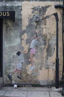 A mural on a weathered wall portrays two children, one reaching upwards and the other appearing to jump. The mural is placed beneath a streetlamp and next to a building sign. The background shows peeling paint and cracked texture, providing a rustic ambiance.