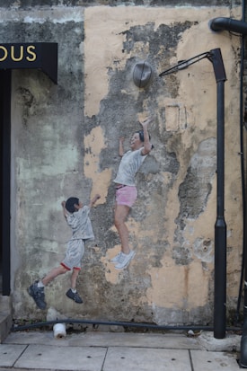 A mural on a weathered wall portrays two children, one reaching upwards and the other appearing to jump. The mural is placed beneath a streetlamp and next to a building sign. The background shows peeling paint and cracked texture, providing a rustic ambiance.