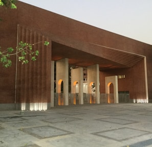 A modern architectural structure featuring a large brick facade with geometric patterns. The building includes tall, vertical openings with arched tops, which are illuminated by warm lights. A few green leaves are visible in the foreground, suggesting nearby vegetation.