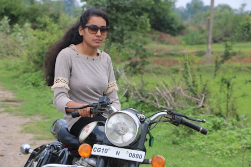 A person wearing sunglasses and a grey sweater with lace detail rides a motorcycle. The setting is outdoors, with lush green foliage and a pathway nearby.