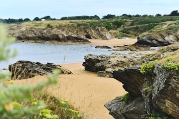 A serene coastal scene with rugged cliffs and a sandy beach. There is calm water lapping against the shore, and the landscape is adorned with patches of lush greenery. A few tents are visible in the distance, suggesting a campsite.