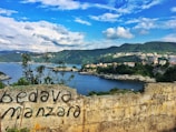 A scenic coastal landscape features a body of water surrounded by lush green hills and scattered buildings. A rustic stone wall in the foreground has graffiti with the words 'Bedava manzara' written on it. The sky is partly cloudy, offering a mix of sunlight and shadow across the landscape.