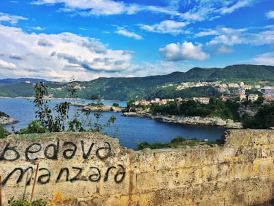 A scenic coastal landscape features a body of water surrounded by lush green hills and scattered buildings. A rustic stone wall in the foreground has graffiti with the words 'Bedava manzara' written on it. The sky is partly cloudy, offering a mix of sunlight and shadow across the landscape.