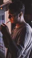 A person is standing indoors, lighting a cigarette. The lighting creates a soft glow on their face, and they seem to be focused on the task. The atmosphere is dimly lit, contributing to a moody, introspective feeling.