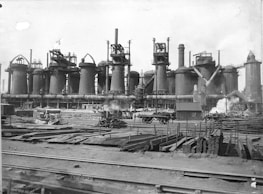 A large industrial facility with multiple tall cylindrical structures, likely blast furnaces, accompanied by numerous pipes and chimneys emitting steam. The foreground contains stacks of metal beams and rails, with scattered construction materials and a railway track.