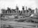 A large industrial facility with multiple tall cylindrical structures, likely blast furnaces, accompanied by numerous pipes and chimneys emitting steam. The foreground contains stacks of metal beams and rails, with scattered construction materials and a railway track.