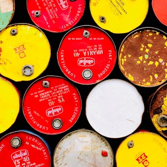 A collection of colorful oil drums arranged closely together. The drums have vibrant red, yellow, white, and brown tops with visible brand logos and labels. The surface of some drums is clean, while others show signs of rust and wear.