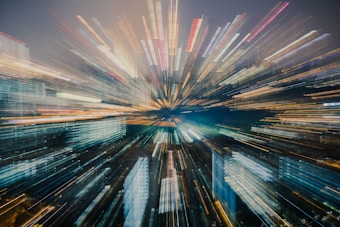 A cityscape at night with buildings illuminated by artificial lights, creating a dynamic zoom effect that centers on a focal point, giving a sense of motion and energy.