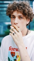 A person with curly hair is smoking a cigarette while wearing multiple rings and a t-shirt with a flower design and the word 'forward' printed on it. The background is blurred, featuring shades of blue and green.