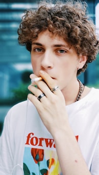 A person with curly hair is smoking a cigarette while wearing multiple rings and a t-shirt with a flower design and the word 'forward' printed on it. The background is blurred, featuring shades of blue and green.