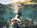 A person is underwater, reaching out toward the camera, surrounded by clear blue water and coral formations. The sunlight creates a ripple effect on the water's surface, adding a dynamic and immersive perspective.