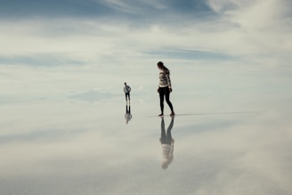 Two people stand on a vast, reflective surface that resembles a mirror, creating an illusion that they are walking in the sky. The horizon is distant and blurred, blending with the cloudy sky above. The atmosphere is serene and otherworldly, with a slight hint of isolation.