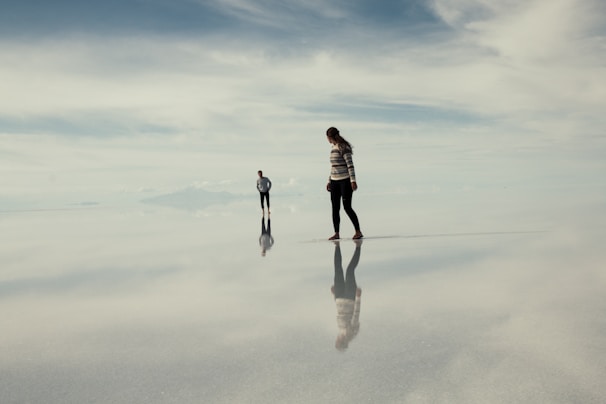 Two people stand on a vast, reflective surface that resembles a mirror, creating an illusion that they are walking in the sky. The horizon is distant and blurred, blending with the cloudy sky above. The atmosphere is serene and otherworldly, with a slight hint of isolation.