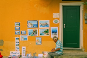 An elderly man is seated on a step next to a vibrant yellow wall adorned with several paintings, showcasing colorful landscapes and seascapes. A green door is situated next to the display of artwork, and the man appears to be engrossed in his work, possibly drawing or reviewing. The wall features peeling paint and a couple of decorative signs.