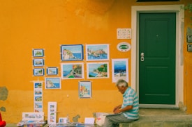 An elderly man is seated on a step next to a vibrant yellow wall adorned with several paintings, showcasing colorful landscapes and seascapes. A green door is situated next to the display of artwork, and the man appears to be engrossed in his work, possibly drawing or reviewing. The wall features peeling paint and a couple of decorative signs.