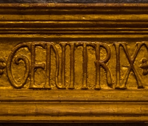 A close-up view of the word 'GENITRIX' carved into a wooden or metallic surface. The letters are raised and have an ornate style, with the background and lettering appearing to be gold.