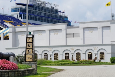 The image displays a sports stadium with a predominantly white exterior. The wall features multiple engravings indicating the graduation years of different classes, such as 'Class of 1958', 'Class of 1960', and others. There is a flagpole with a flag, manicured greenery, and flower bushes near a cannon on the left side of the image. A stone monument with the year 1926 inscribed is also visible. The seating area of the stadium shows blue and yellow seats, along with various flags on the top structure.