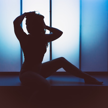 A silhouette of a person sitting on a surface with one knee bent and one arm raised to their head. The background features a soft gradient of blue light, creating a serene and artistic atmosphere.